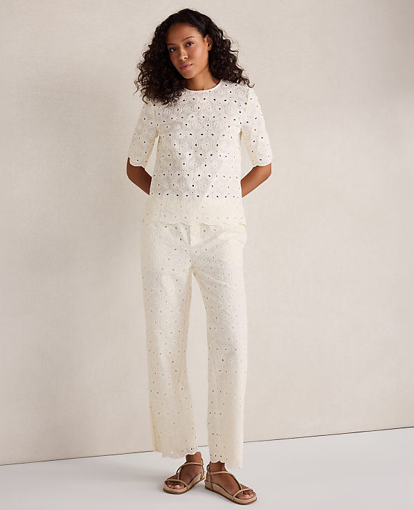 A woman in a white eyelet top and matching pants stands against a plain, light-colored background.