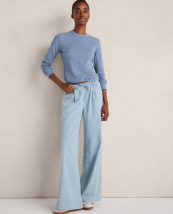 A woman in a blue long-sleeved shirt and wide-leg light blue jeans stands against a plain, textured beige background.