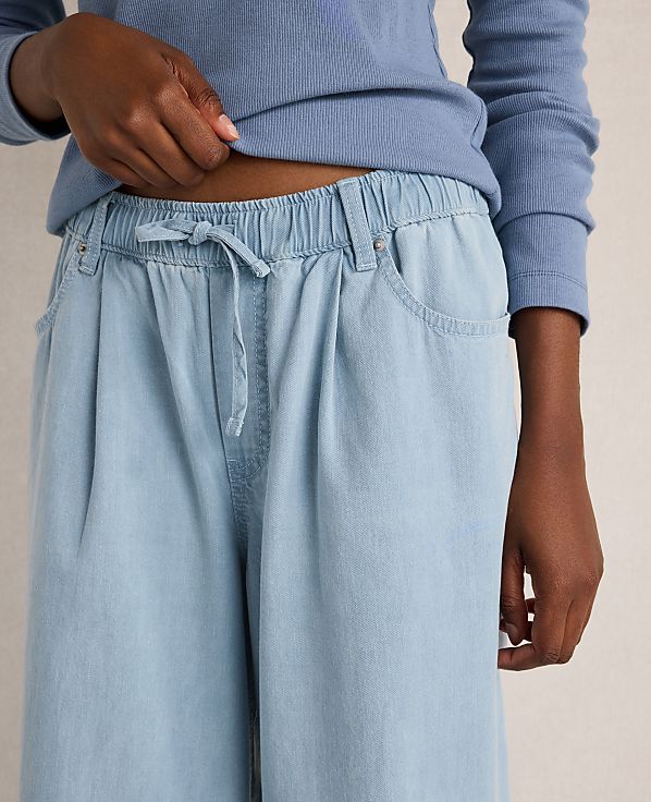 A person's hands adjust the drawstring waist of light blue, wide-leg pants against a plain background.