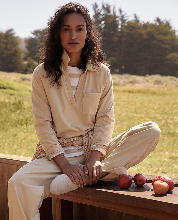 A woman in casual attire sits on a wooden railing with apples in the foreground, while a blurred, natural landscape with trees forms the background.