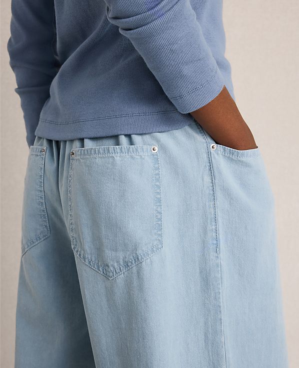 A person in a blue long-sleeved shirt has their hands in the pockets of light blue wide-leg pants against a plain background.