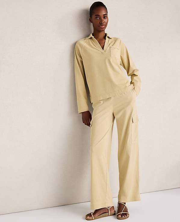 A woman in a light yellow collared shirt and matching wide-leg pants stands against a textured cream wall.
