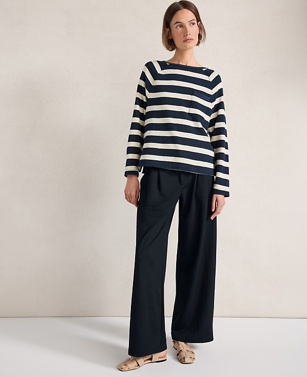 A woman in a striped sweater and wide-leg pants stands against a plain, textured wall.
