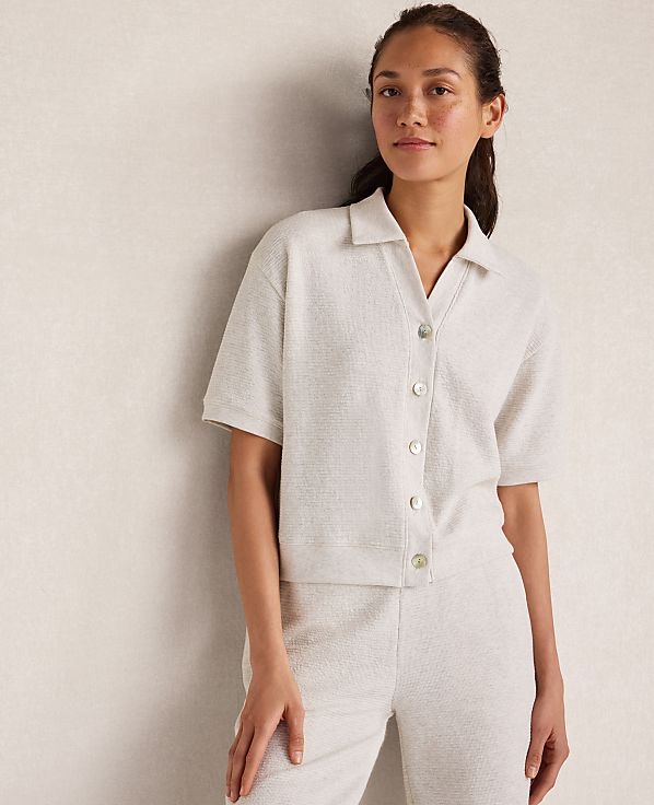 A woman in a cream-colored collared shirt and pants stands against a textured, light-colored wall.