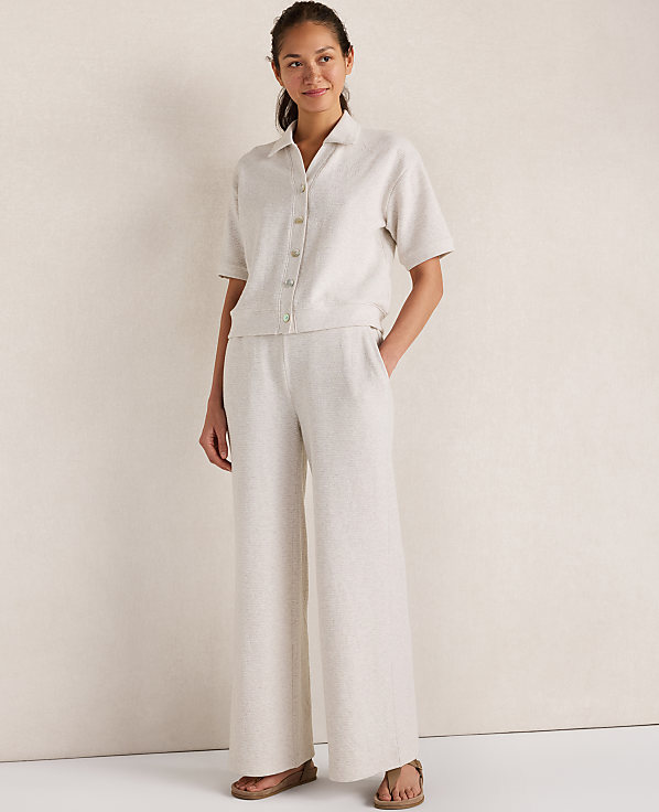A woman in a linen shirt and wide-leg pants stands against a textured, light-colored wall.