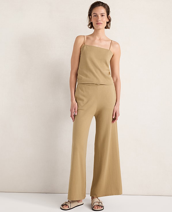 A woman in a tan knit camisole and wide-leg pants stands against a plain, textured cream background.