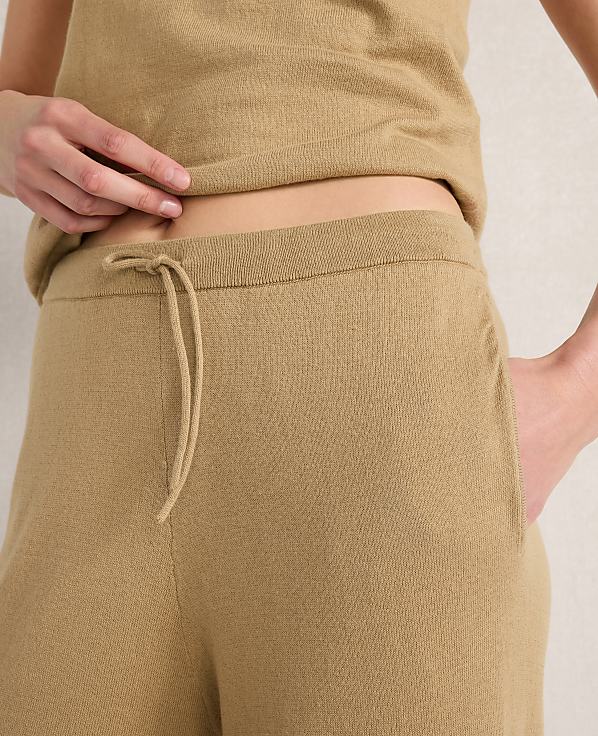 A person's midsection is shown in the foreground, wearing tan loungewear with a drawstring waist, against a plain, light-colored background.