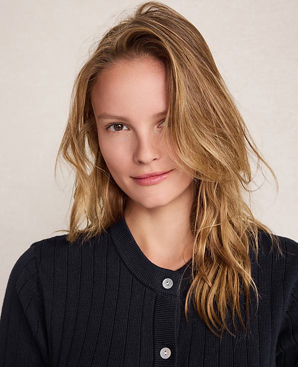A young woman with blonde hair and a dark blue cardigan smiles gently against a plain, light-colored background.