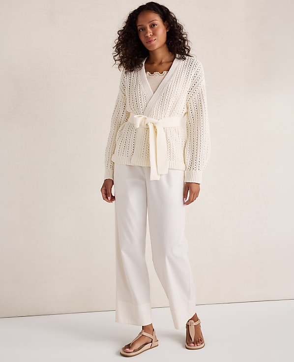A woman in a cream-colored wrap cardigan and wide-leg pants stands against a plain, light-colored background.