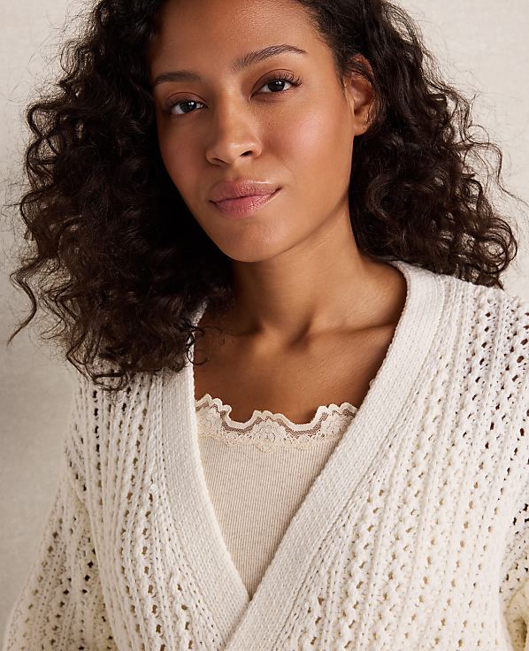 A woman with curly dark hair wears a cream-colored, open-knit cardigan over a cream-colored top with a scalloped neckline, against a soft, blurred background.