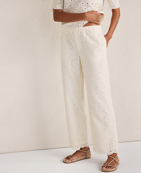 A person stands in cream eyelet pants and sandals against a plain, light-colored background.