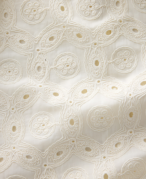 Intricate white eyelet embroidery forms a repeating geometric pattern against a soft, textured fabric background.