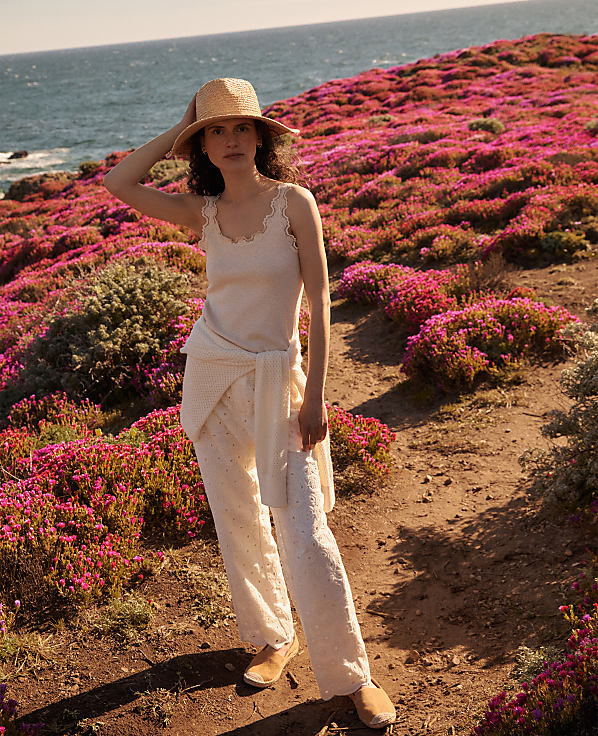 A woman in a straw hat and white outfit stands on a dirt path surrounded by vibrant pink wildflowers, with the ocean visible in the background.