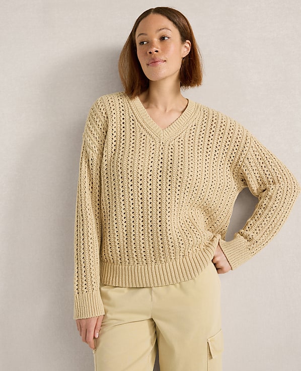 A woman in a beige v-neck sweater and light-colored pants stands against a textured gray wall.