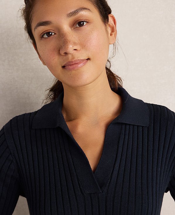 A young woman with dark hair pulled back smiles gently in a dark blue ribbed polo shirt against a plain, light-colored background.