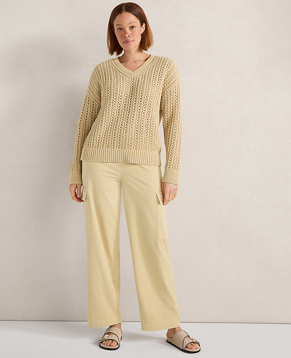 A woman in a beige v-neck sweater and light yellow cargo pants stands against a textured, light-colored wall.