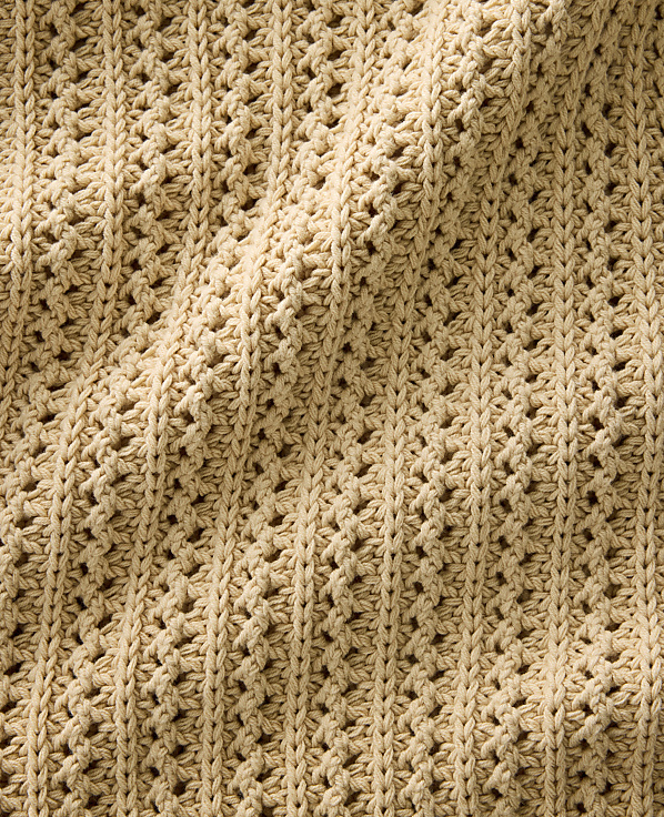 A textured, beige crocheted fabric with a repeating openwork pattern is draped and folded.