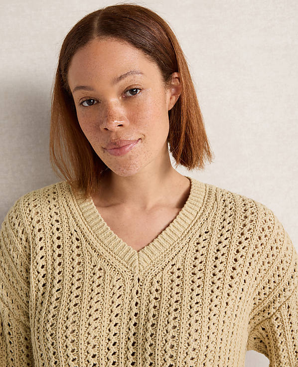 A woman with short brown hair and freckles smiles gently in a light yellow, open-knit sweater against a textured, light-colored background.