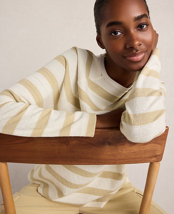 A young woman in a striped sweater leans on a wooden chair against a plain background.