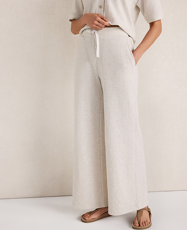 A person wearing light-colored wide-leg pants and sandals stands against a textured, neutral background.