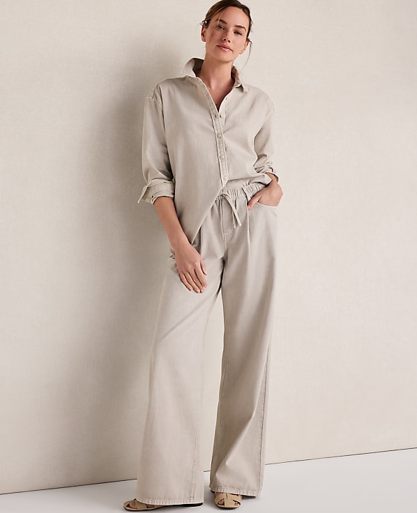 A woman in a beige linen shirt and wide-leg pants stands against a plain, textured wall.
