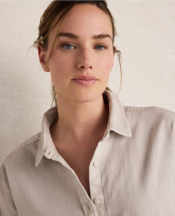 A woman with light skin and blue eyes, wearing a light-colored button-down shirt, is captured in a close-up shot against a textured, neutral background.