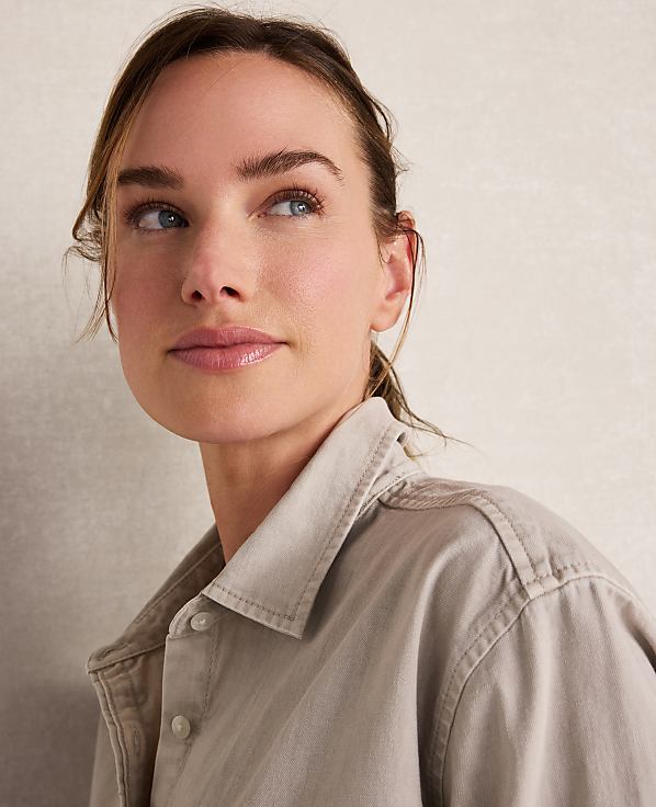 A woman with light skin and brown hair pulled back looks up and to the left, wearing a light brown collared shirt against a plain, textured background.