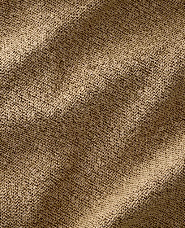 A close-up view of a textured, tan knitted fabric fills the frame, with soft shadows and highlights creating depth across its surface.