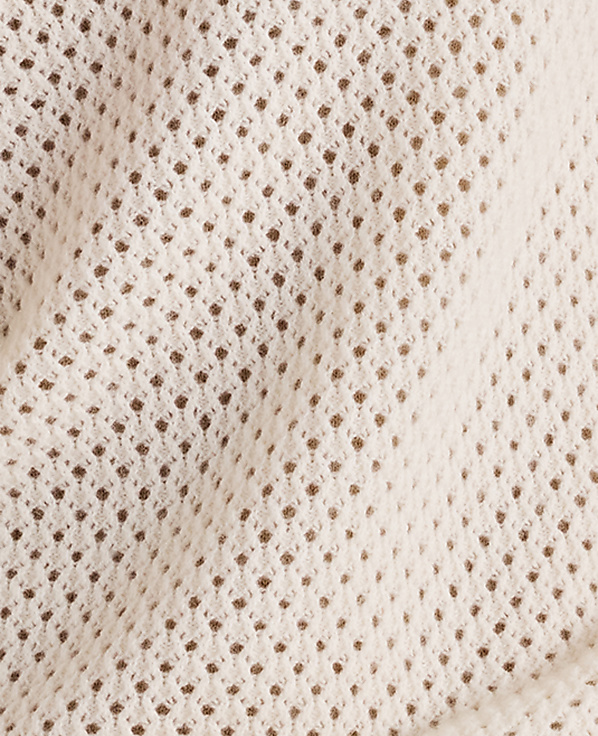 A close-up view of a cream-colored, loosely woven fabric with a textured, open-knit pattern fills the frame, revealing subtle shadows and folds in the material.