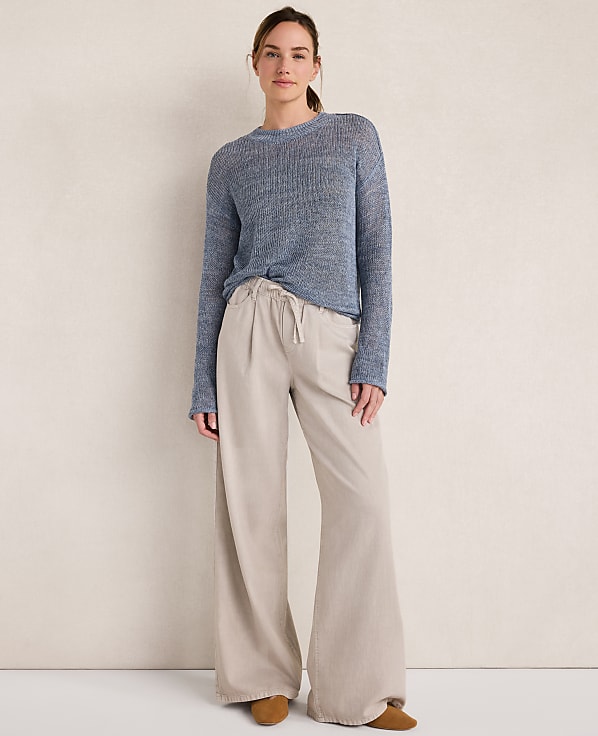 A woman in a blue sweater and beige pants stands against a textured cream wall.