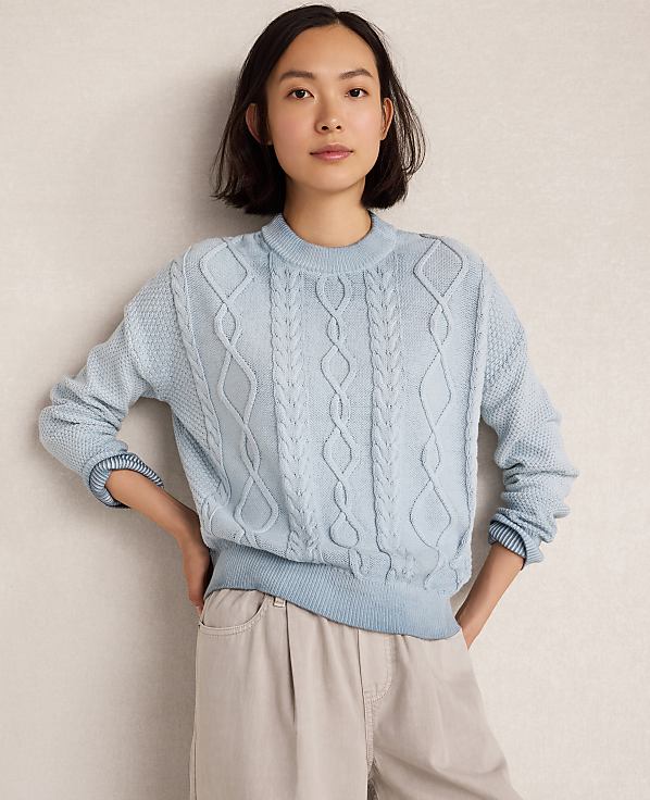 A young woman in a light blue cable-knit sweater and beige pants stands against a plain, light-colored wall.