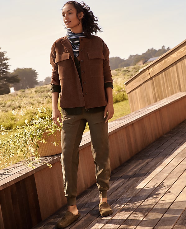 A woman stands on a wooden deck in the foreground, wearing a brown jacket and green pants, with a grassy hillside and trees in the background.