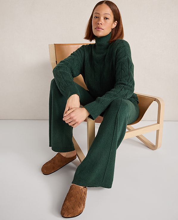 A woman in a green cable-knit turtleneck and matching pants sits on a modern chair with brown leather accents, against a plain, light-colored wall.