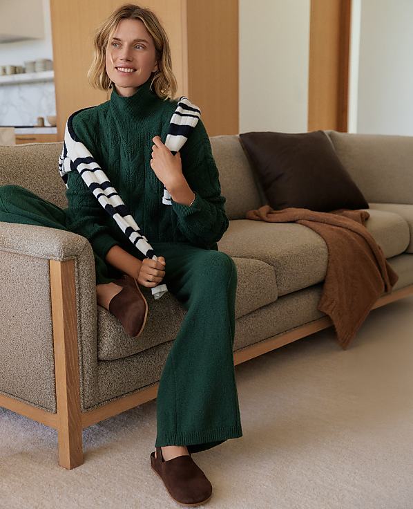 A woman in a green sweater and pants sits on a couch with a striped scarf draped over her shoulder, while a blurred background shows a modern living room.