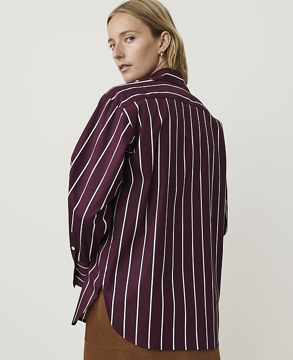 A woman in a burgundy and white striped shirt stands against a plain, light background.