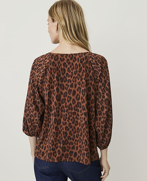 A woman in a brown leopard-print top and dark jeans stands against a plain white background.