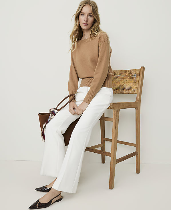 A woman in a camel sweater and white pants sits on a wooden stool with a woven seat, against a plain white background.