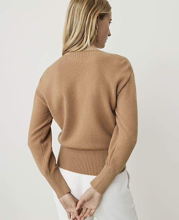 A woman in a tan sweater and white pants stands with her back to the camera against a plain white background.