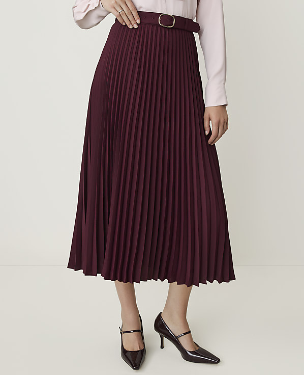 A burgundy pleated midi skirt with a gold buckle belt is worn with a light pink blouse and dark patent heels against a plain white background.