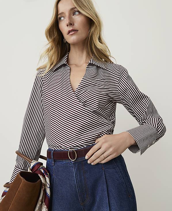 A woman in a striped wrap top and denim jeans stands against a plain background, holding a brown bag.