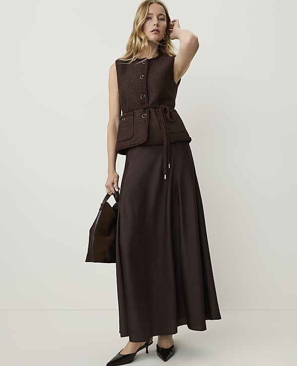 A woman in a brown vest and skirt stands against a plain white background, holding a dark brown handbag.