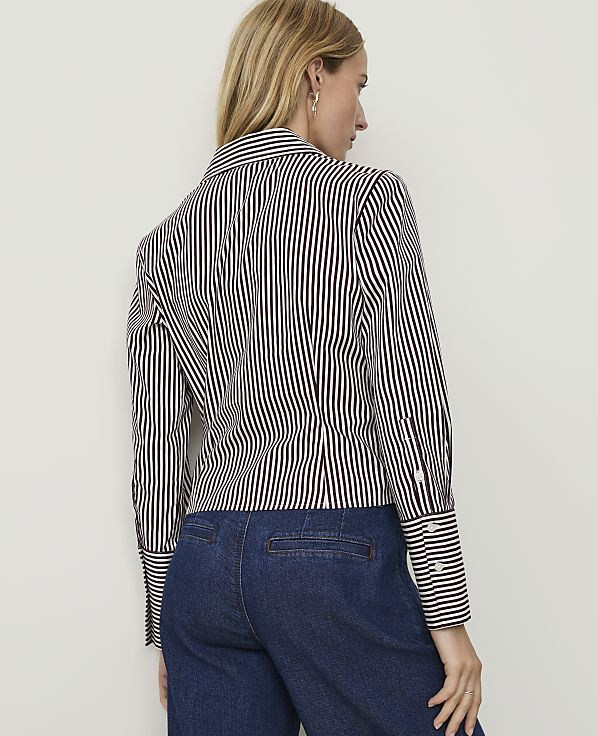 A woman in a striped blouse and denim pants stands against a plain wall.