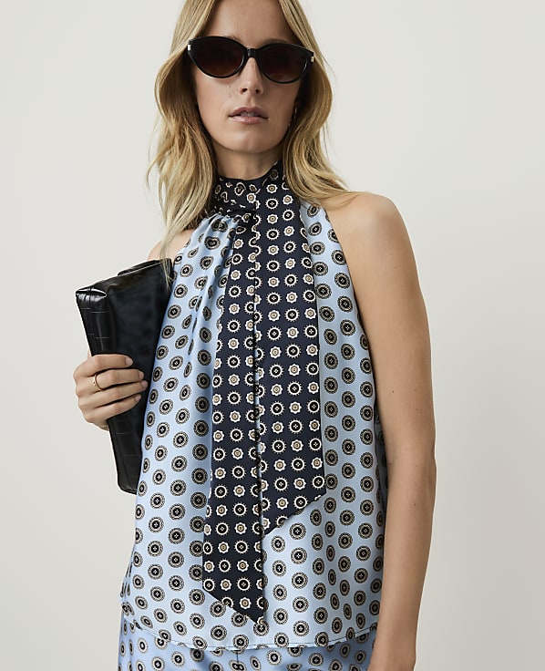 A woman in a light blue patterned halter top and sunglasses holds a black clutch in front of a plain background.