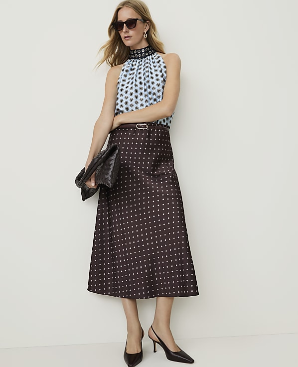 A woman in a patterned top and skirt stands against a plain white background, holding a clutch.