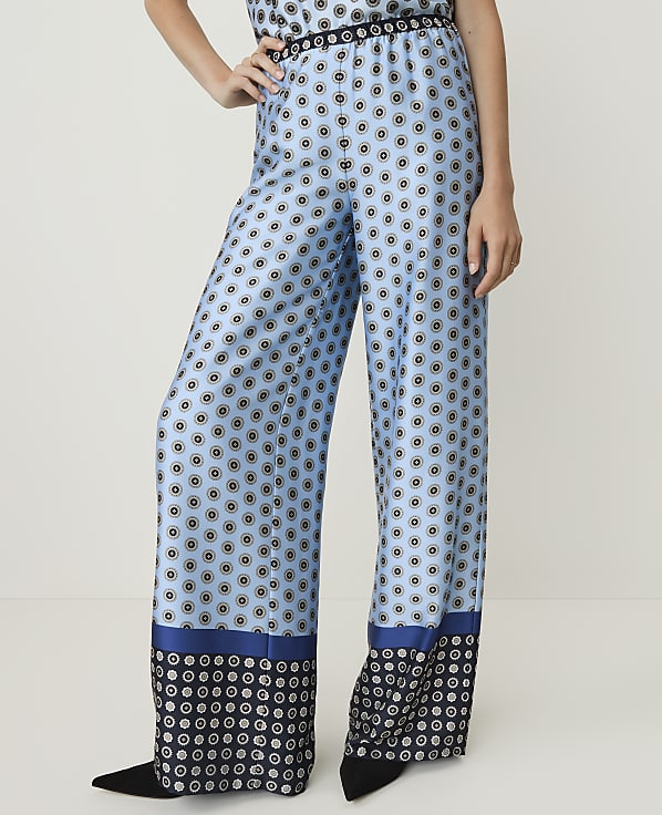 A person stands in wide-leg, light blue patterned pants with a dark blue and black geometric border at the hem, against a plain white background.