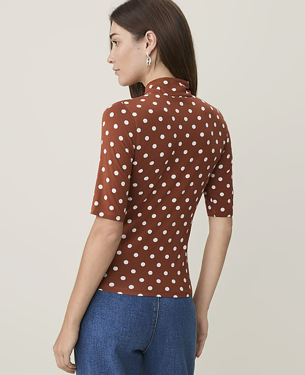A woman in a brown polka dot shirt and blue jeans stands against a plain, light-colored background.
