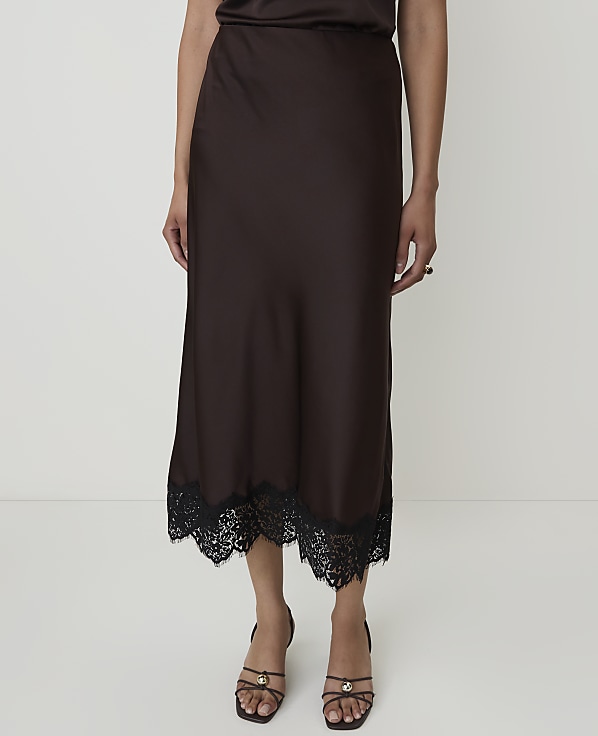 A person stands in front of a plain white background, wearing a brown satin skirt with a black lace trim and strappy sandals.