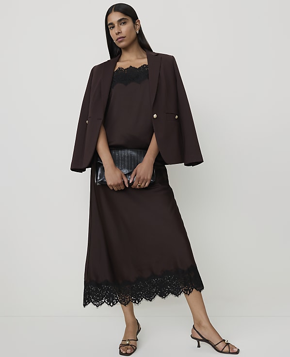 A woman in a brown slip dress and blazer holds a black clutch in front of a plain white background.