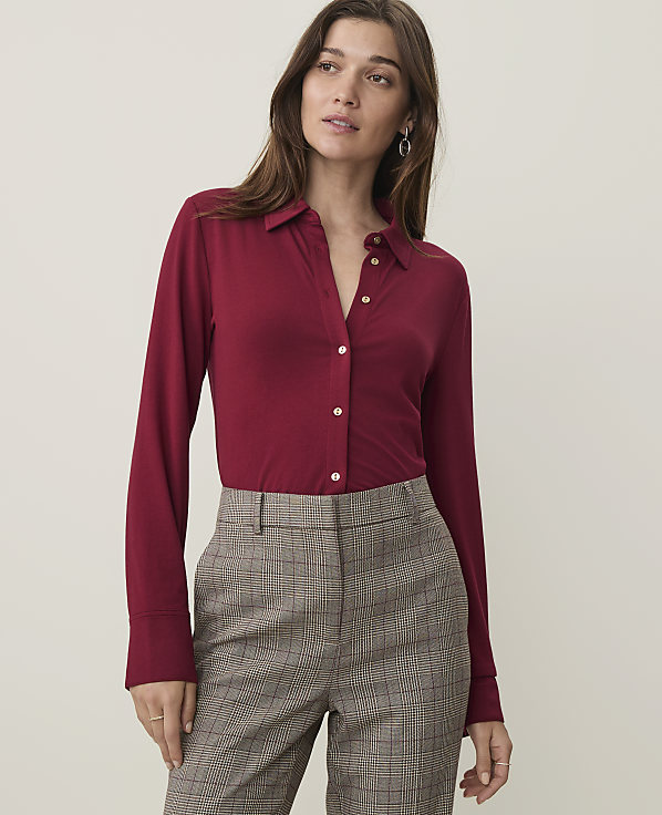 A woman in a maroon button-up shirt and plaid pants stands against a plain, light-colored background.