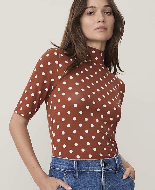 A woman in a brown polka dot turtleneck and blue jeans stands against a plain, light-colored background.